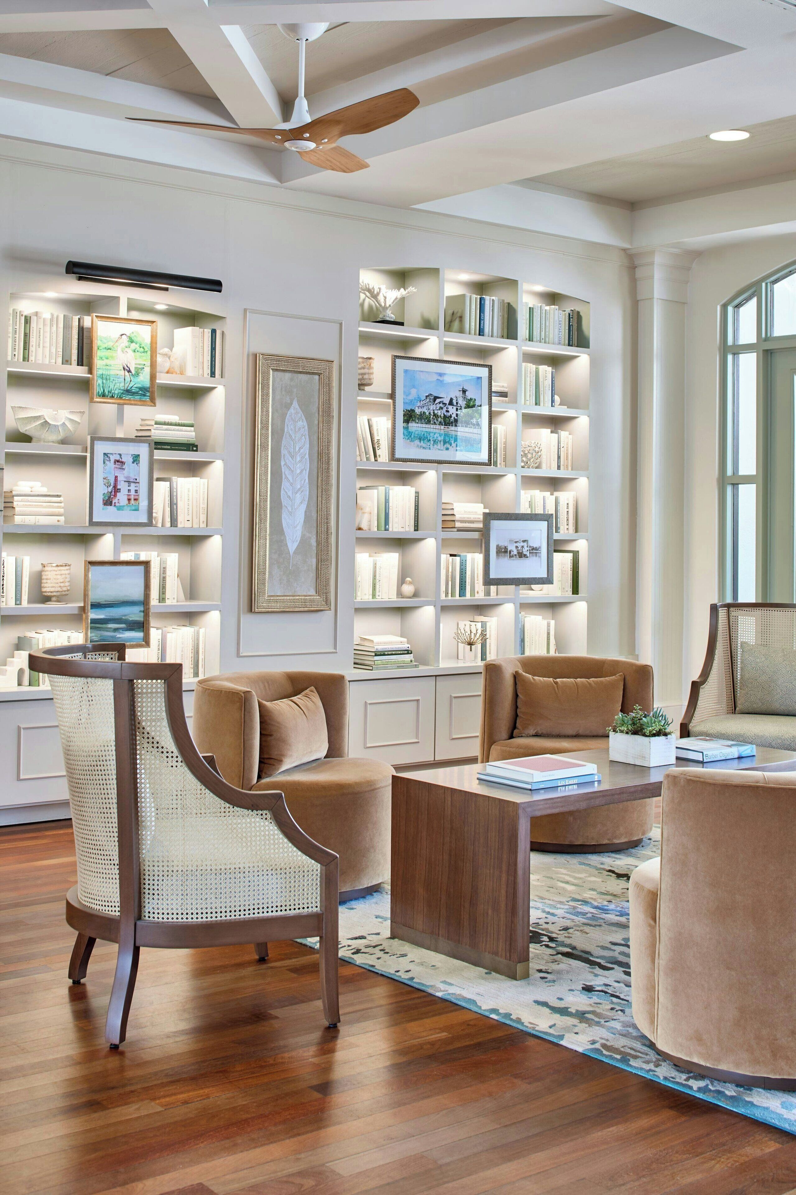 A spacious living room with a large bookshelf, two armchairs, a coffee table, and a ceiling fan.