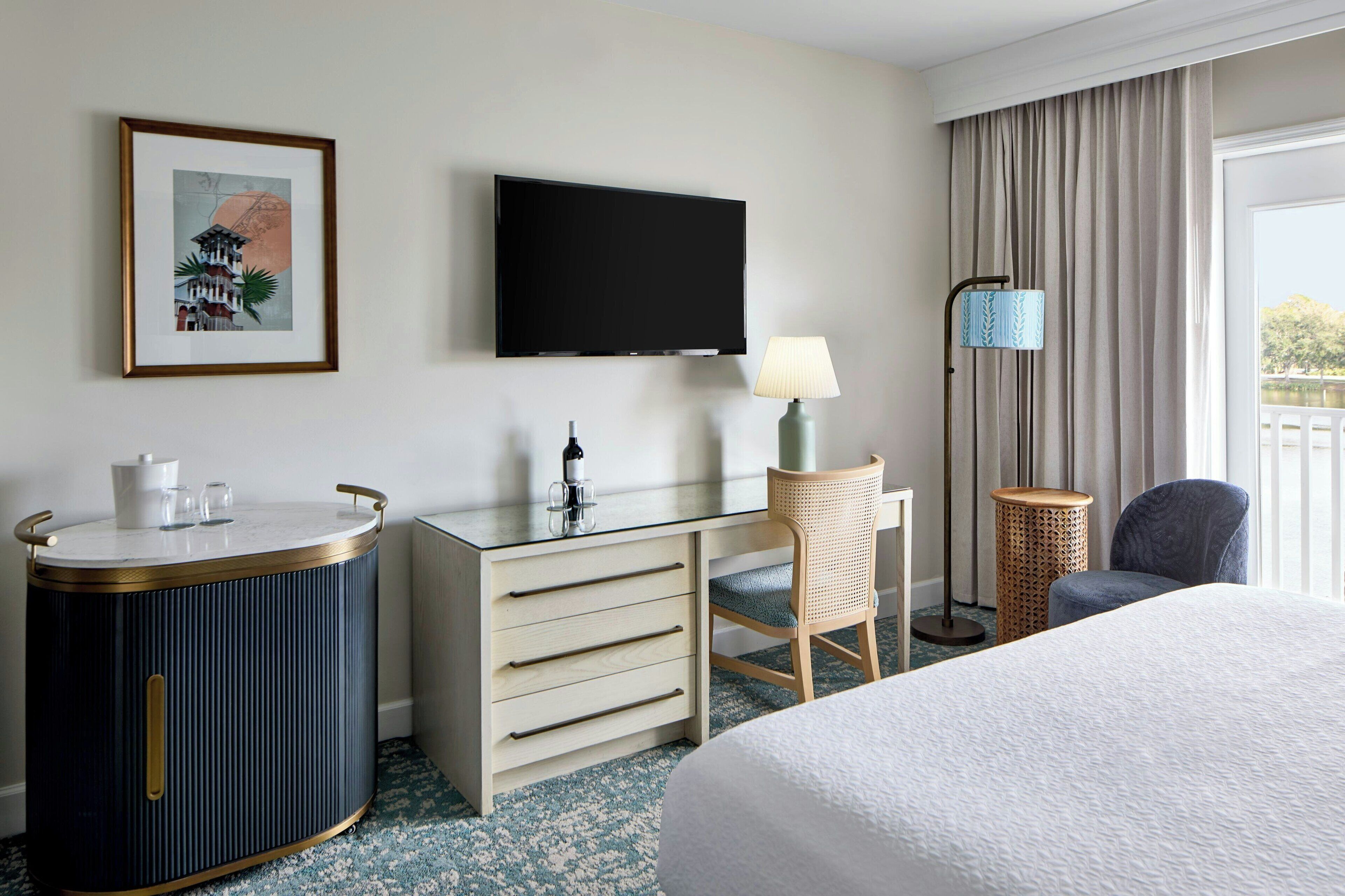A hotel room with a bed, a desk with drawers, a television, a lamp, a chair, and a framed picture on the wall.