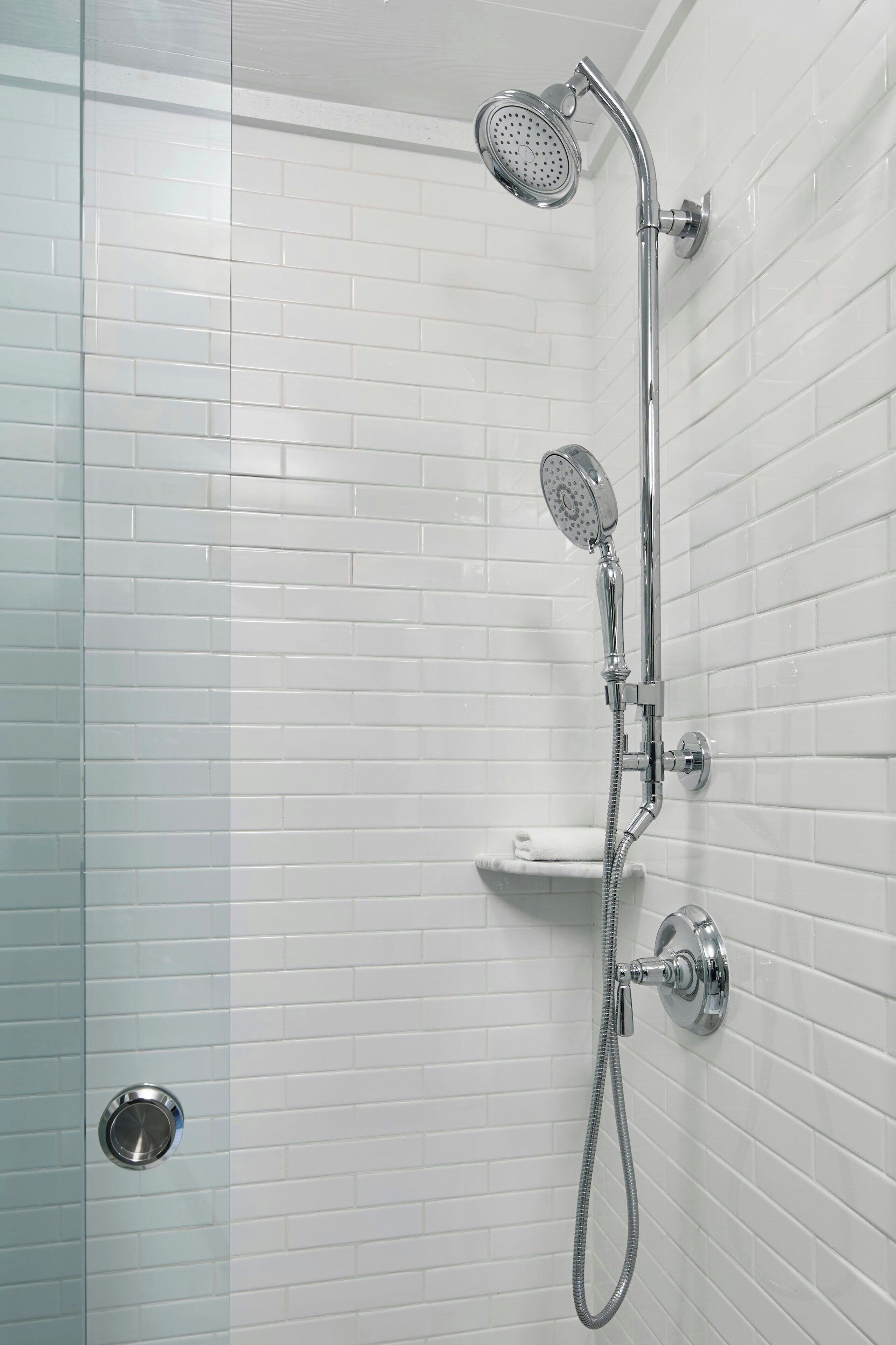 Een douche met een handdouche en een vaste douchekop, bevestigd aan een wand met witte tegels.