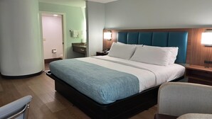 Una camera d'albergo con un letto grande, un comodino in legno, una sedia e un bagno visibile attraverso una porta aperta.