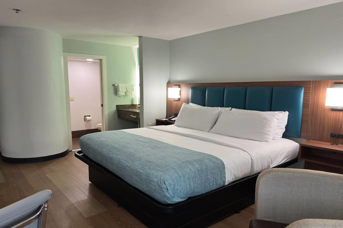A hotel room with a large bed, a wooden nightstand, a chair, and a bathroom visible through an open door.