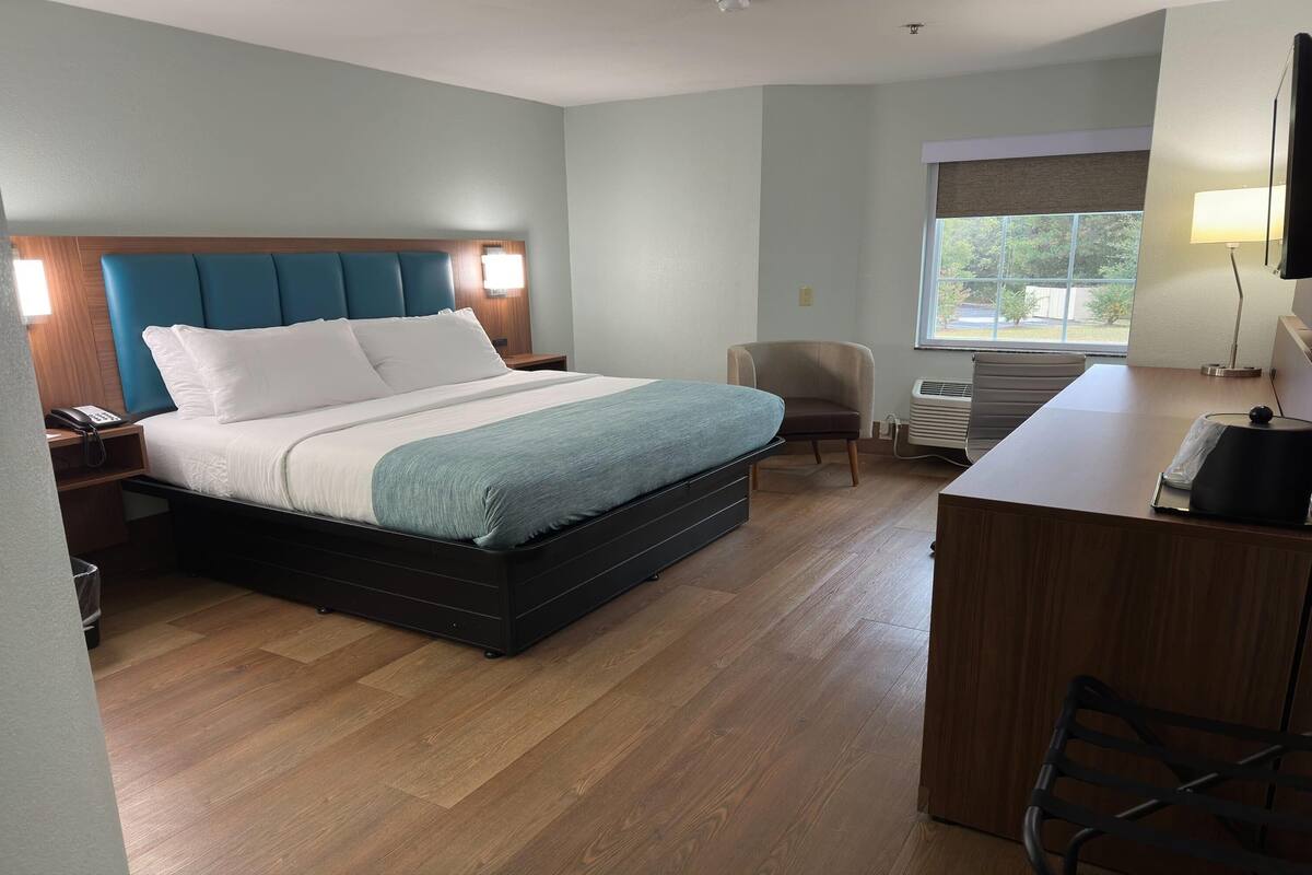 A hotel room with a bed, bedside table, chair, desk, and window.