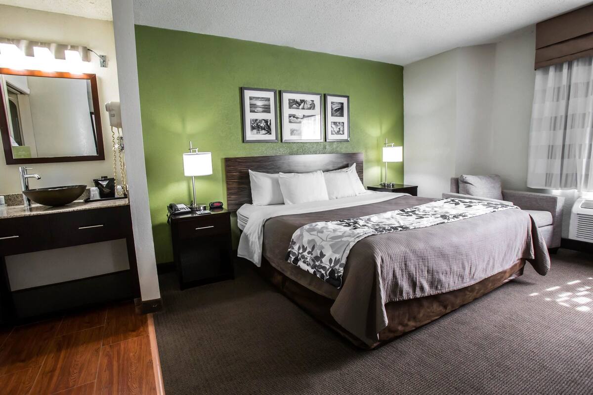 A hotel room with a bed, bedside tables, a vanity with a mirror, a sink, and framed pictures on the wall.