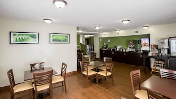 Zona pranzo con tavoli e sedie in legno, angolo cottura con frigorifero e macchina del caffè, e quadri con immagini di felci alle pareti.