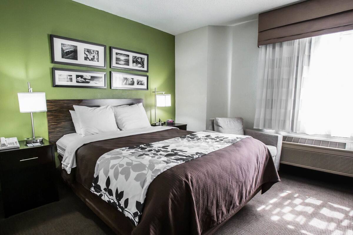 A hotel room with a bed, bedside tables, a lamp, a chair, a window with curtains, and framed pictures on the wall.