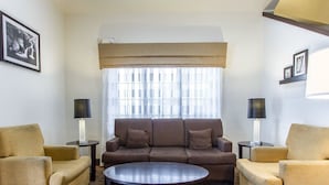 Een moderne woonkamer met een bruine bank, twee fauteuils en een ronde salontafel. Er staan twee lampen op bijzettafels, een groot raam met dunne gordijnen en ingelijste foto’s aan de muur.