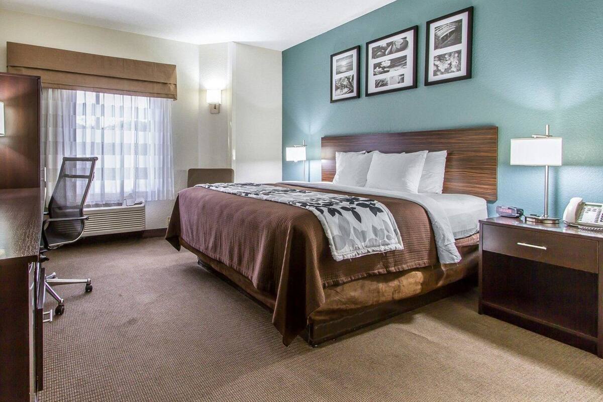 Quarto de hotel com uma cama grande, uma escrivaninha com luminária, uma cadeira e uma janela com cortinas.