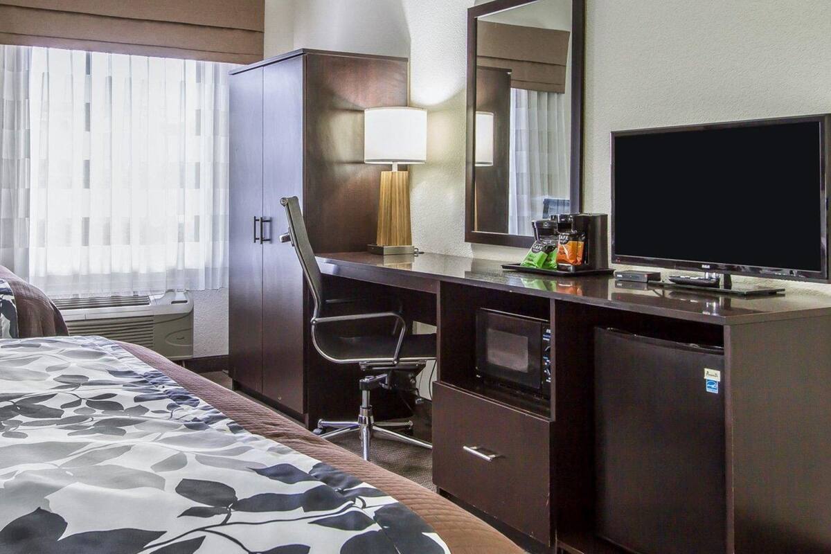 Quarto de hotel com cama, escrivaninha, cadeira, televisão, cafeteira e espelho.