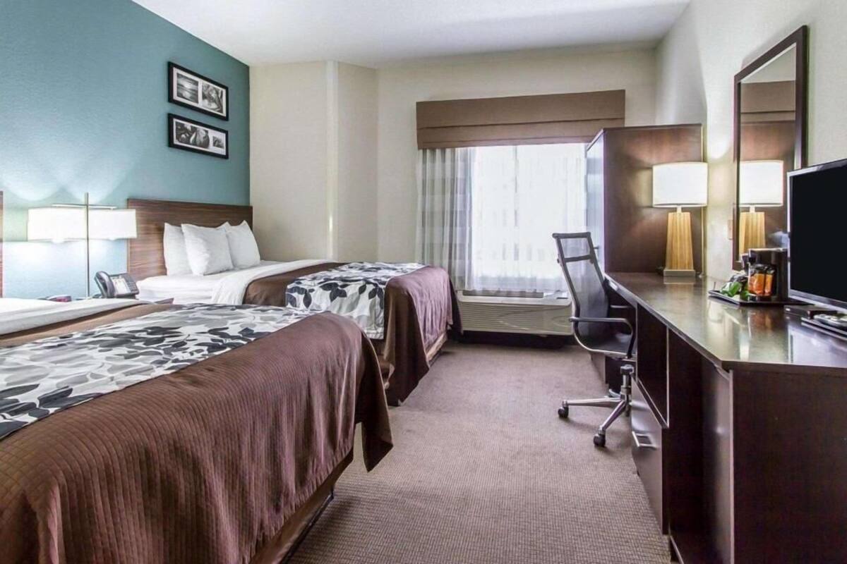 Quarto de hotel com duas camas, uma escrivaninha, uma cadeira, uma televisão e uma janela com cortinas.