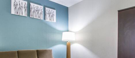 A hotel room with a beige sofa, a small wooden table, and a floor lamp. There are three wall-mounted artworks depicting plants.