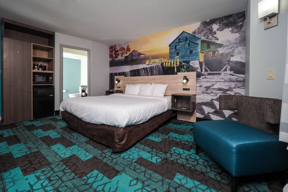 A hotel room with a large bed, a wooden headboard, a nightstand with a lamp, a blue bench, and a wall mural.