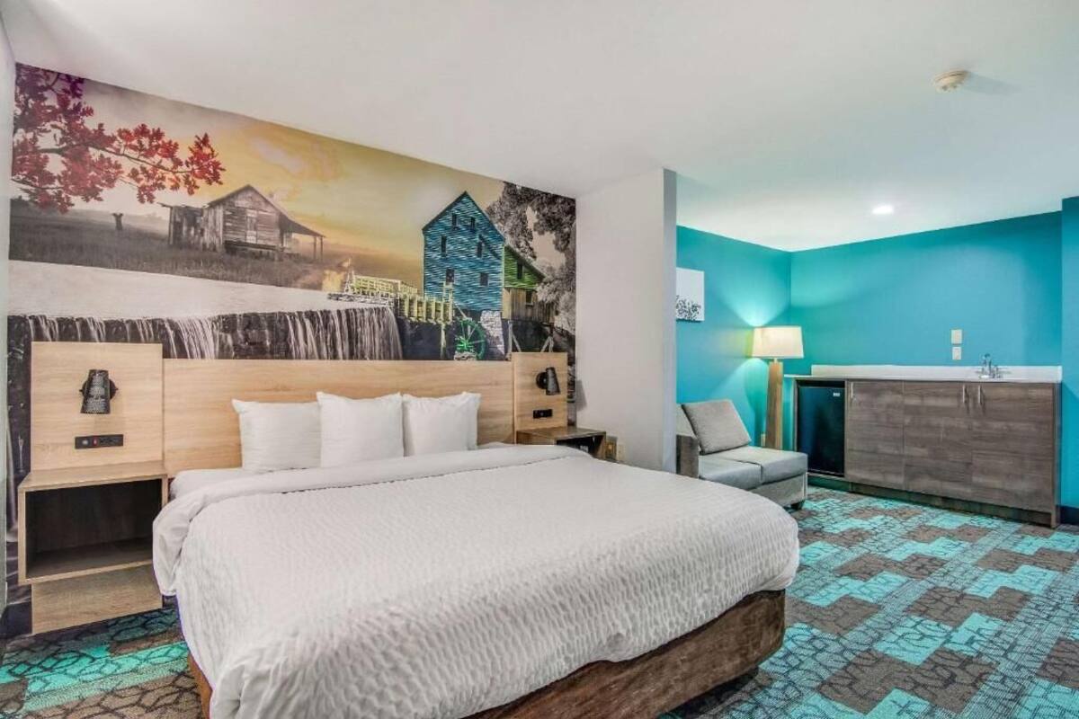 A hotel room with a large bed, a wooden headboard, a wall mural, a grey armchair, a small desk, a lamp, a sink, and a patterned rug.