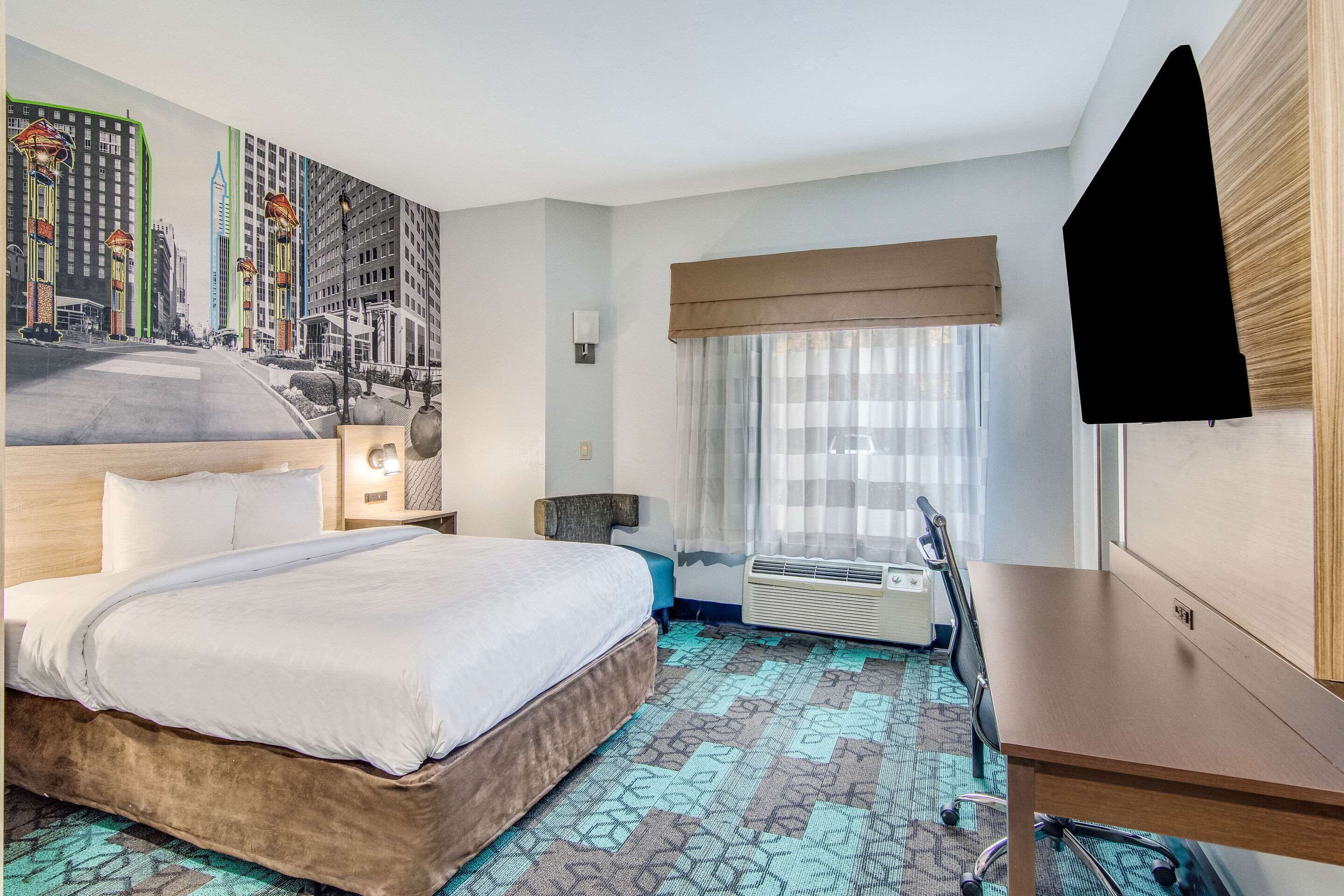 Habitación de hotel con cama, escritorio, silla, ventana con cortinas, televisión montada en la pared y alfombra estampada.