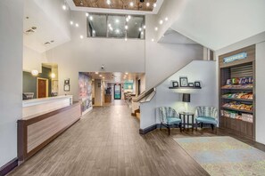 A hotel lobby with a reception desk, seating area, staircase, and snack bar. - Clarion Pointe Wake Forest - Raleigh North (Wake Forest)