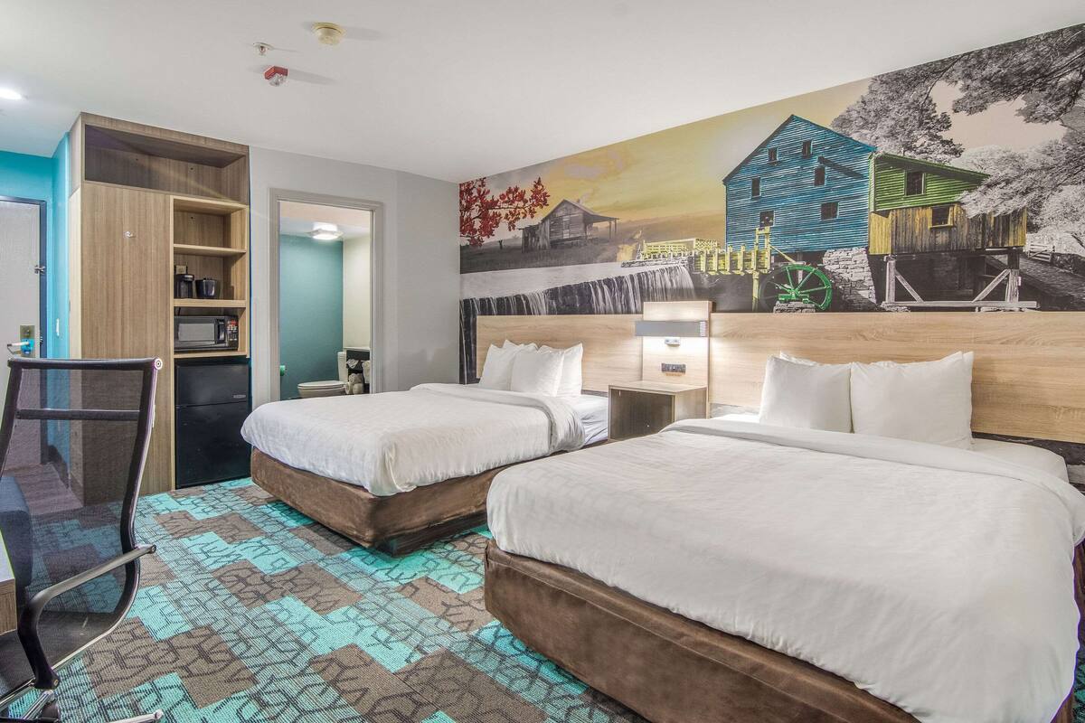 A hotel room with two beds, a desk, a chair, a microwave, and a shelf with items.
