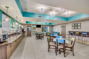 A spacious cafeteria with multiple tables and chairs, a counter with a microwave, and a television screen.