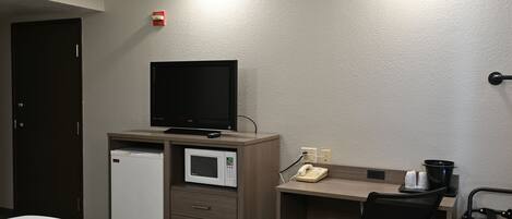 A hotel room with a bed, a desk with a computer monitor, a microwave, a telephone, and a trash bin.