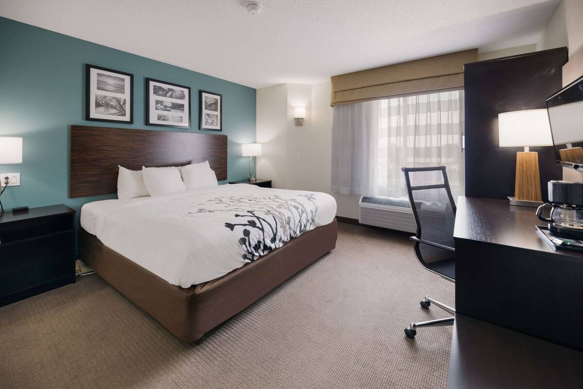 A hotel room with a large bed, a desk with a chair, a window with curtains, and wall-mounted lamps.