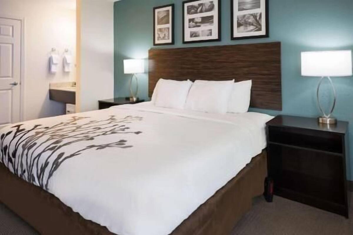 A hotel room with a large bed, two bedside tables, a nightstand, a wall-mounted lamp, and a framed photo collage.