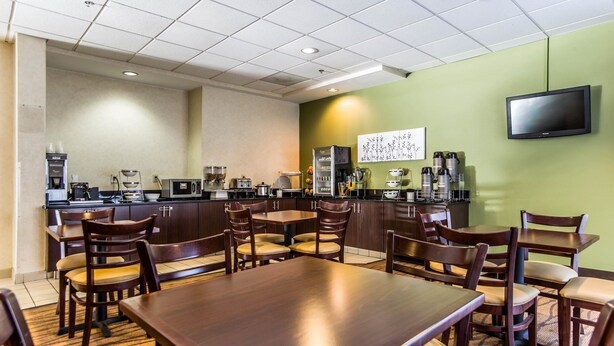 A hotel dining area with wooden tables and chairs, a kitchen in the background, and a flat-screen TV on the wall. - Sleep Inn Columbia Airport (West Columbia)