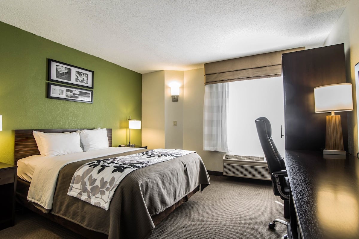 A hotel room with a bed, bedside lamps, a desk with a chair, a window with curtains, and wall-mounted pictures.
