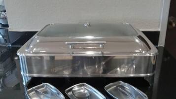 Disposição de buffet com recipiente de plástico transparente e utensílios de servir sobre uma mesa preta.
