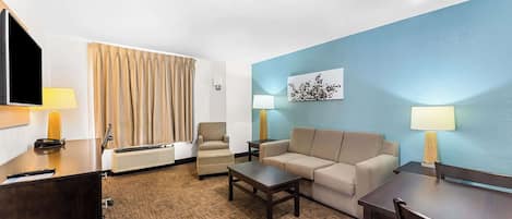 Een hotelkamer met een bureau, twee stoelen, een bank, een salontafel, een televisie en een groot raam met gordijnen.