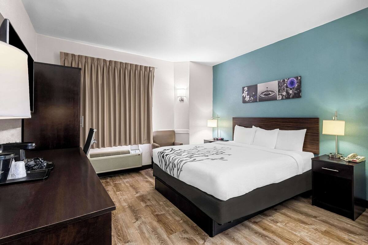 A hotel room with a bed, bedside tables, a desk, a chair, a window with curtains, and artwork on the wall.