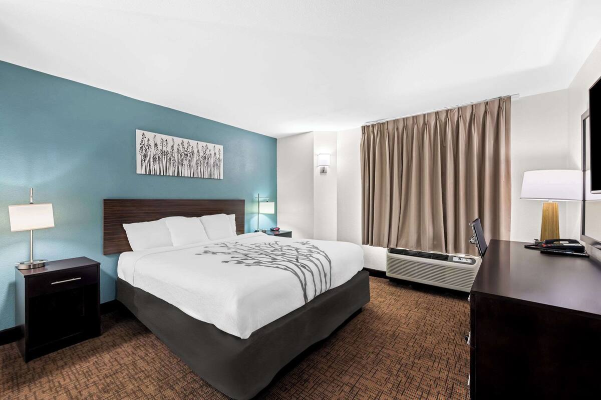 A hotel room with a bed, a nightstand, a desk, a lamp, a wall art piece, a window with curtains, and a wall-mounted air conditioning unit.