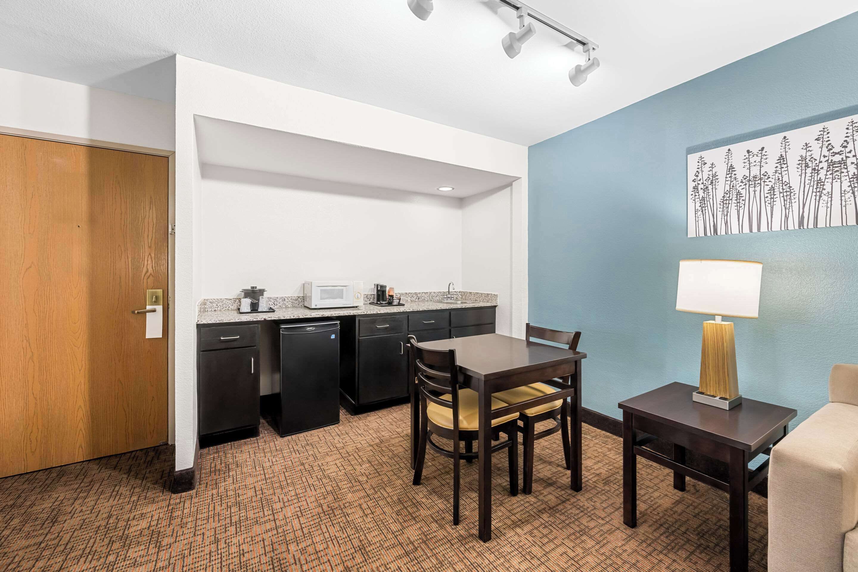A compact hotel room with a kitchenette, a dining area with a table and chairs, a lamp, and a wall-mounted artwork.