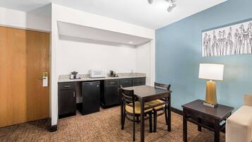 A compact hotel room with a kitchenette, a dining area with a table and chairs, a lamp, and a wall-mounted artwork.