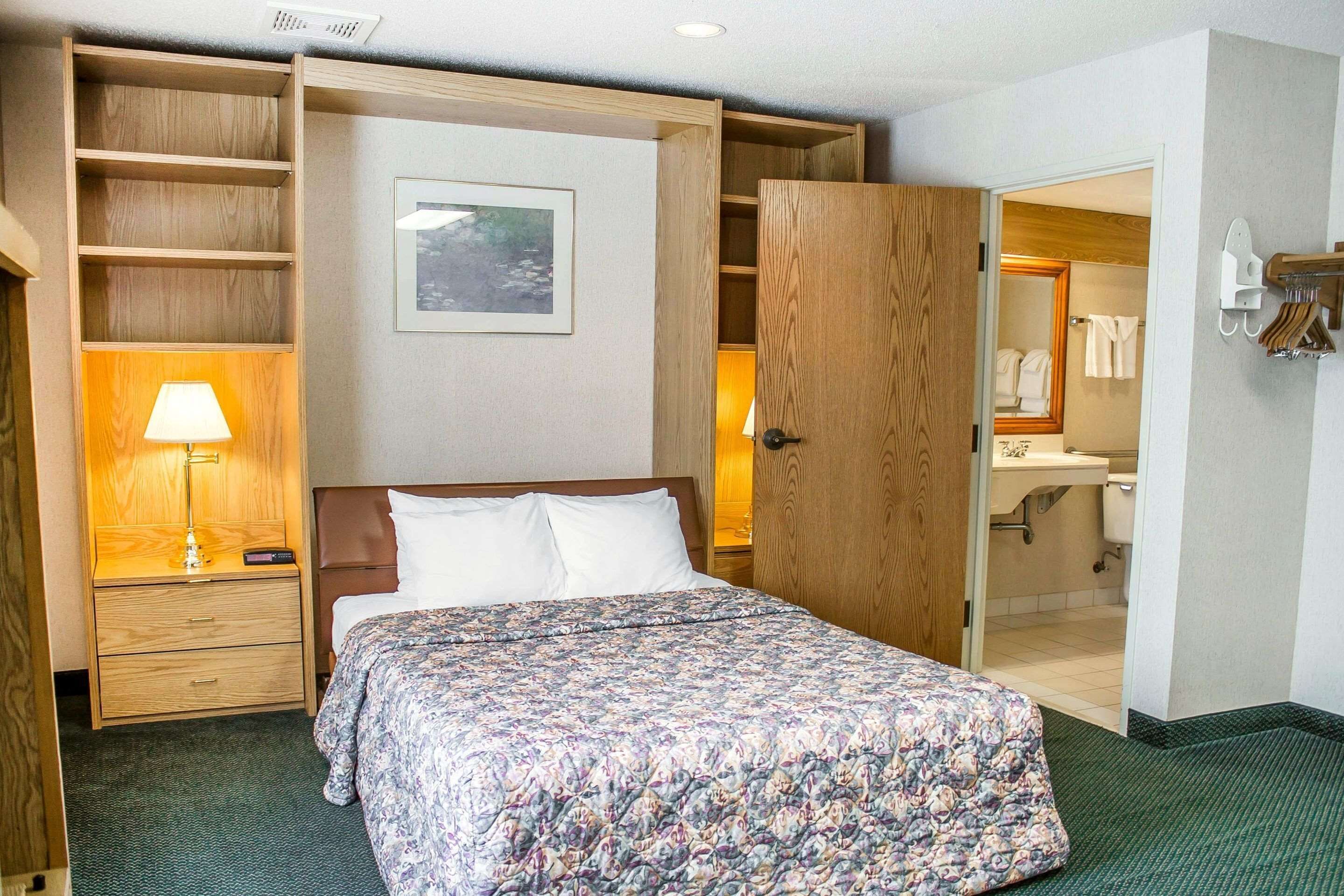 Habitación de hotel con cama, mesita de noche con lámpara, armario de madera y baño visible a través de una puerta abierta.
