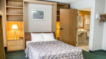 Habitación de hotel con cama, mesita de noche con lámpara, armario de madera y baño visible a través de una puerta abierta.