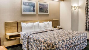 A hotel room with a large bed, wooden headboard, two bedside tables, a nightstand, and a wall-mounted lamp. - Rodeway Inn Abbotsford (Colby)