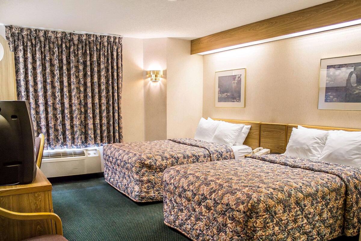 Quarto de hotel com duas camas, uma escrivaninha, uma cadeira, um ar condicionado e uma cortina.