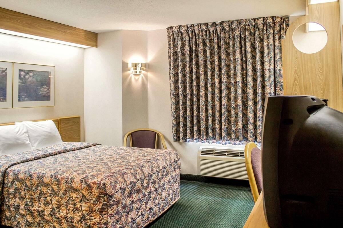 Quarto de hotel com cama, poltrona, televisão, luminária fixada na parede e janela com cortinas.