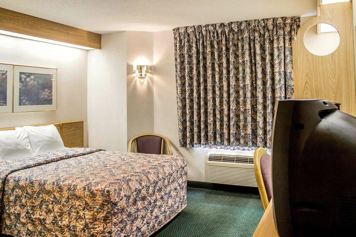 Quarto de hotel com cama, poltrona, escrivaninha com computador, luminária na parede e janela com cortinas.