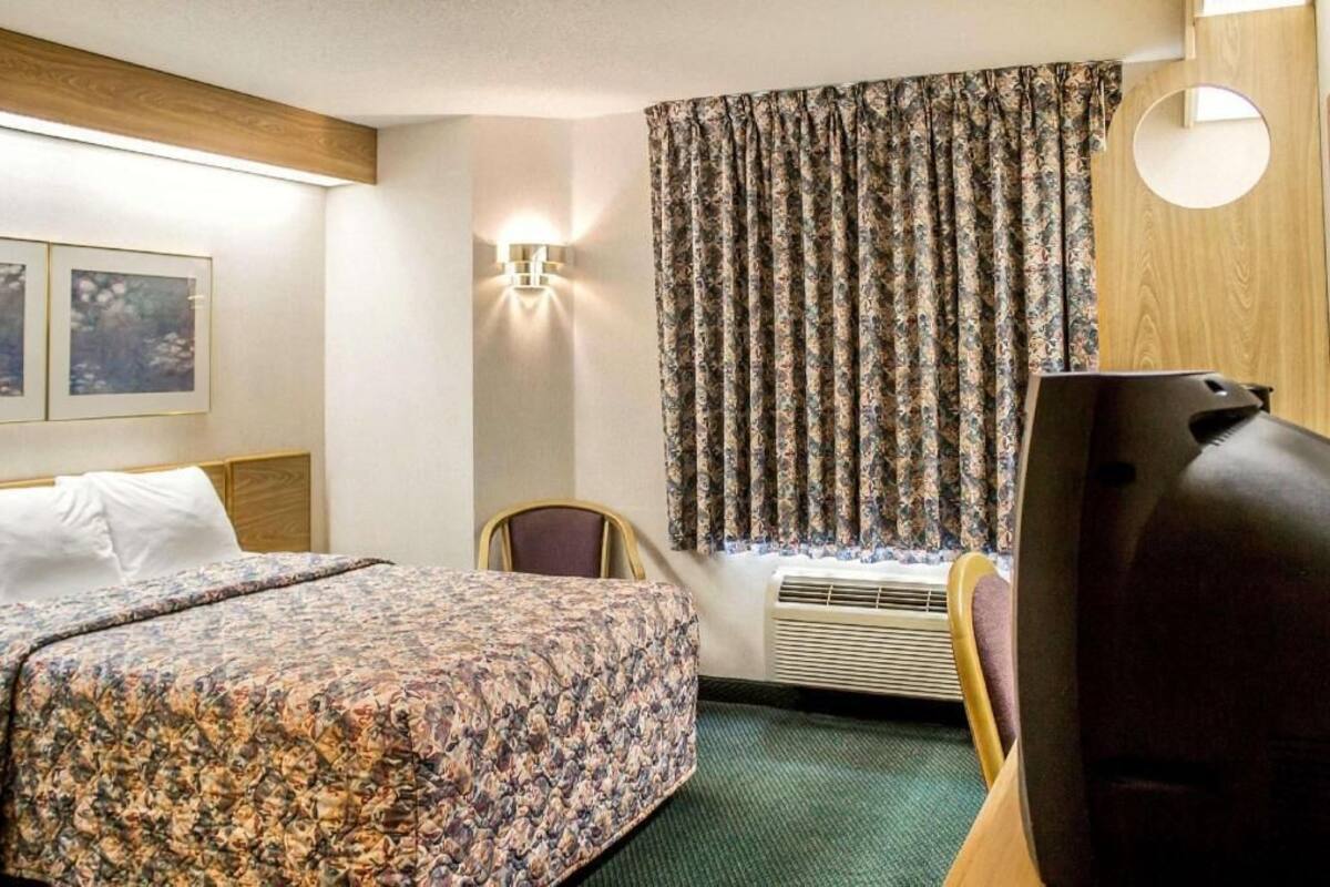 Quarto de hotel com cama, poltrona, televisão, luminária fixada na parede e janela com cortinas.