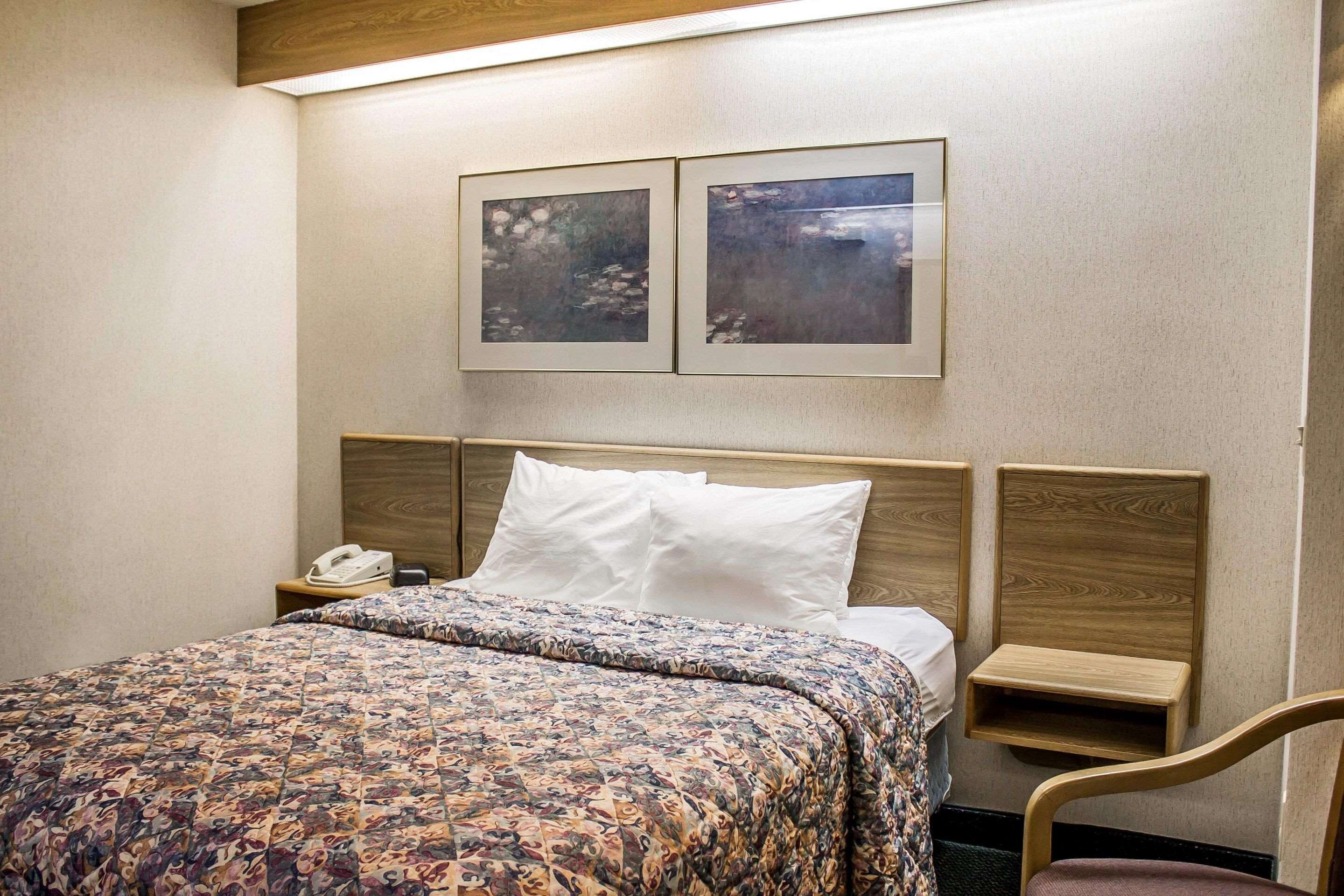 Une chambre d’hôtel avec une tête de lit en bois, une couvre-lit à motifs, une table de chevet avec un téléphone et deux tableaux encadrés au mur.