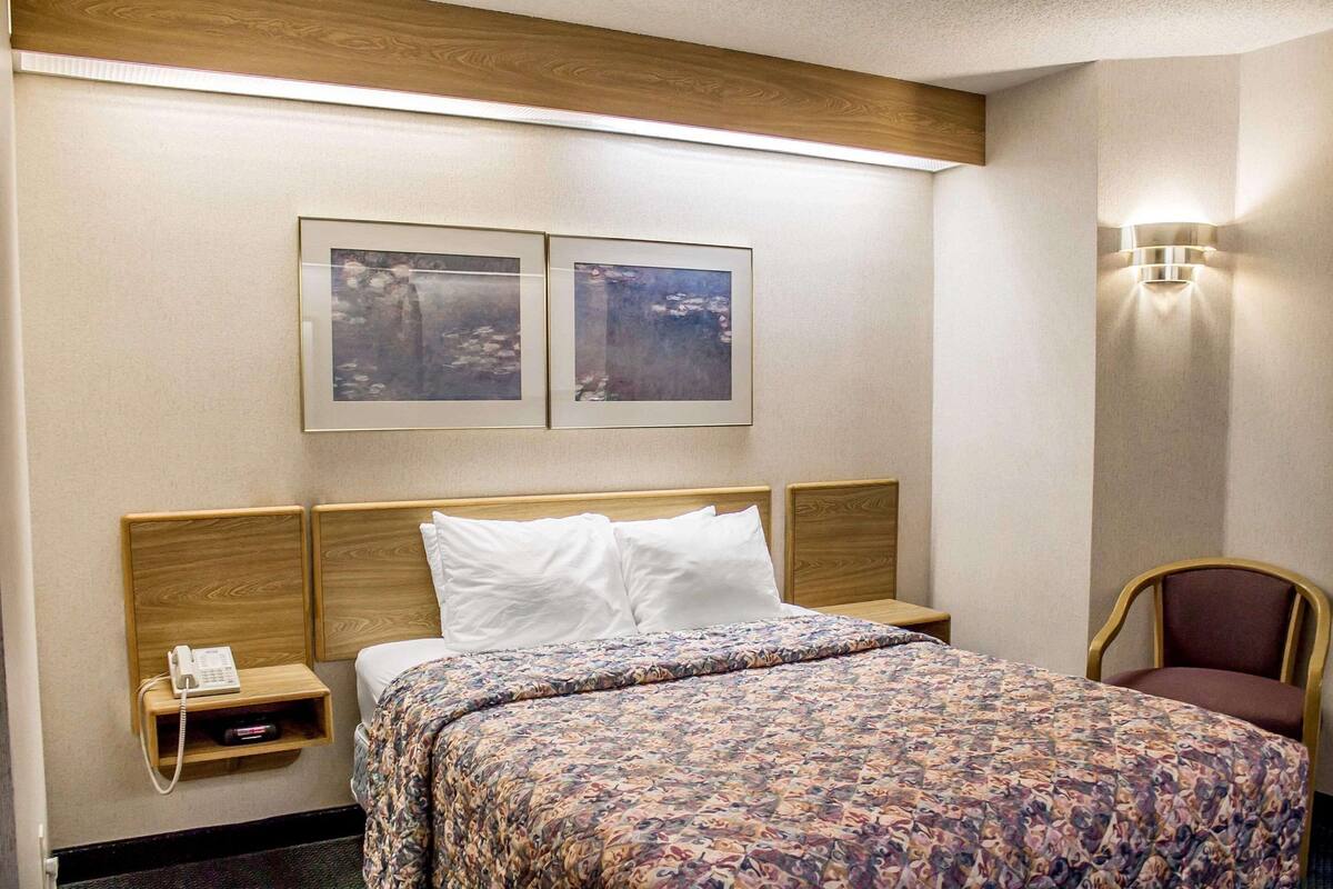 Quarto de hotel com cabeceira de madeira, cama com colcha estampada, duas quadros na parede, telefone de apoio e uma cadeira.