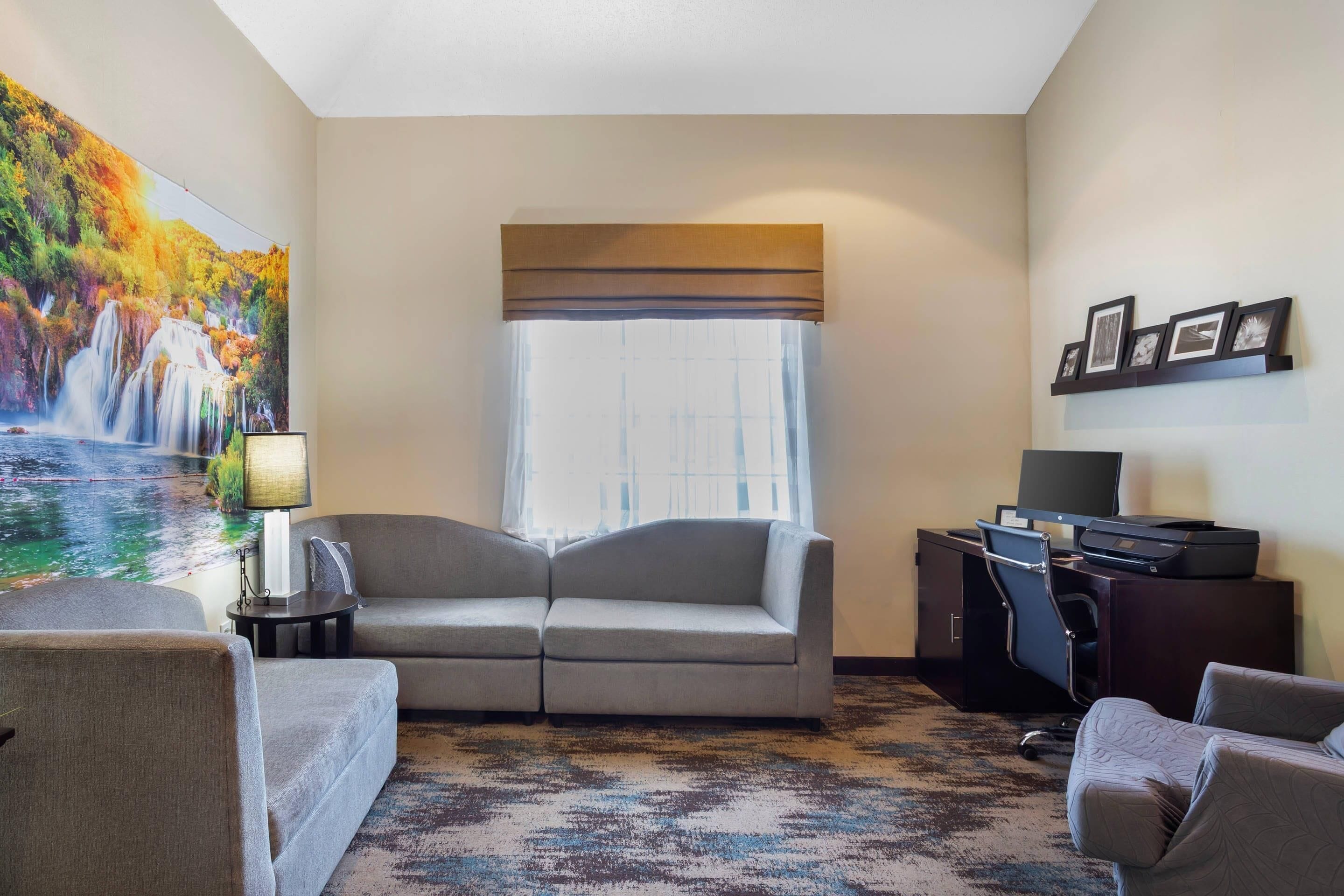 Habitación de hotel con sofá, escritorio con computadora, televisor, ventana con cortinas y un cuadro de una cascada.
