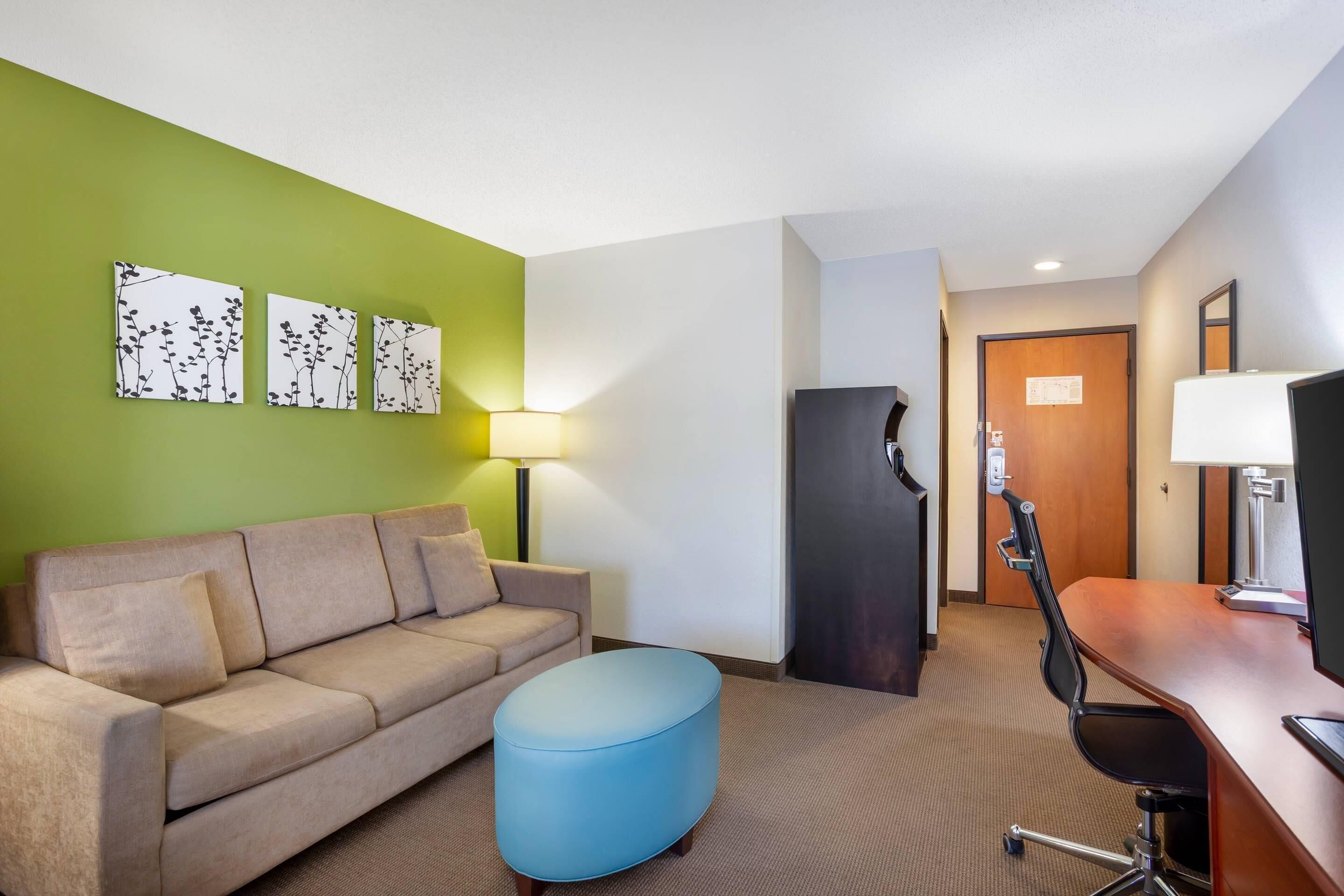 A hotel room with a sofa, a blue ottoman, a desk with a chair, a refrigerator, a lamp, and three wall-mounted artworks.