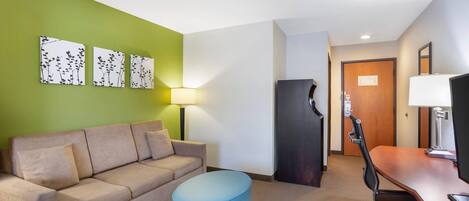 A hotel room with a sofa, a blue ottoman, a desk with a chair, a refrigerator, a lamp, and three wall-mounted artworks.