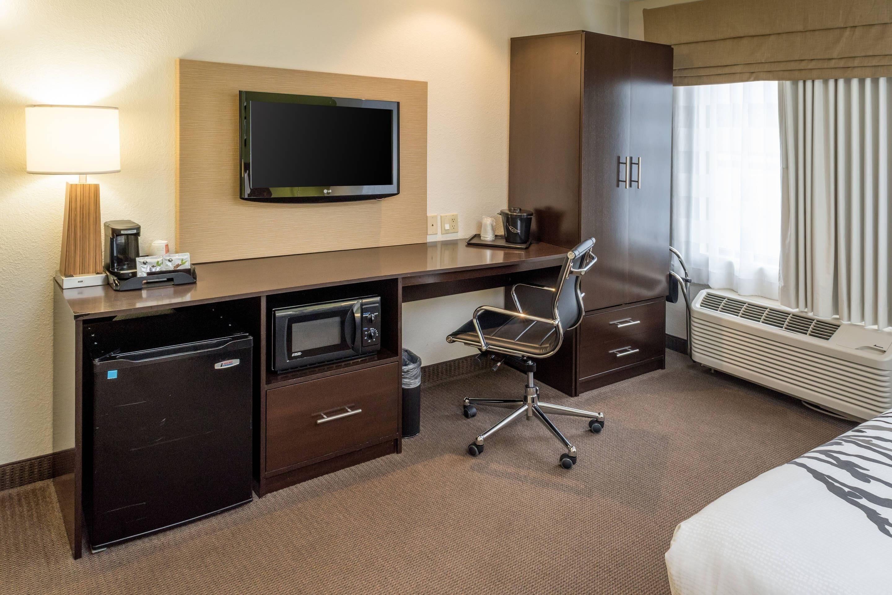 A hotel room with a desk, microwave, mini fridge, TV, air conditioning unit, and a bed.