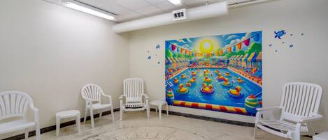 A room with a mural of a sunny outdoor scene, white plastic chairs, and a tiled floor.