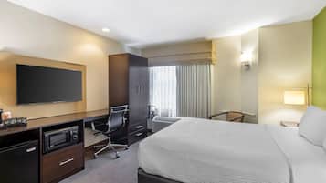 Habitación de hotel con una cama, un escritorio con microondas, un televisor, una silla y una ventana con cortinas.