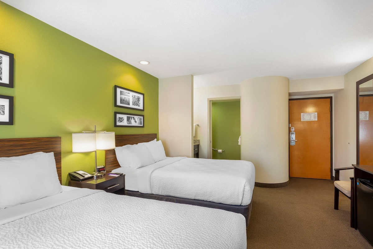 Une chambre d’hôtel avec deux lits, un bureau avec une lampe, une chaise, une porte et des tableaux encadrés au mur.