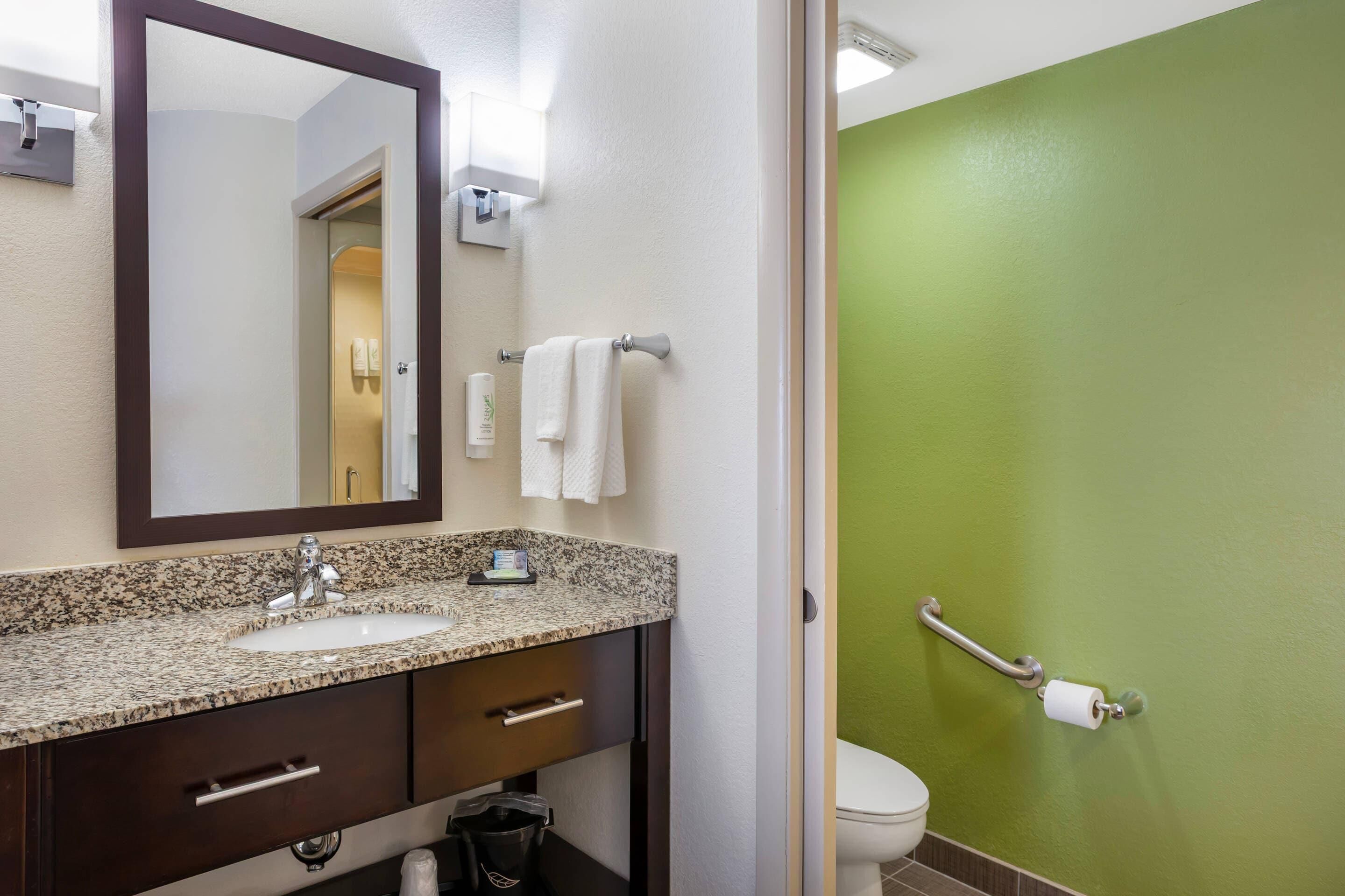 A bathroom with a granite countertop sink, a mirror, a towel rack, and a toilet with a grab bar.
