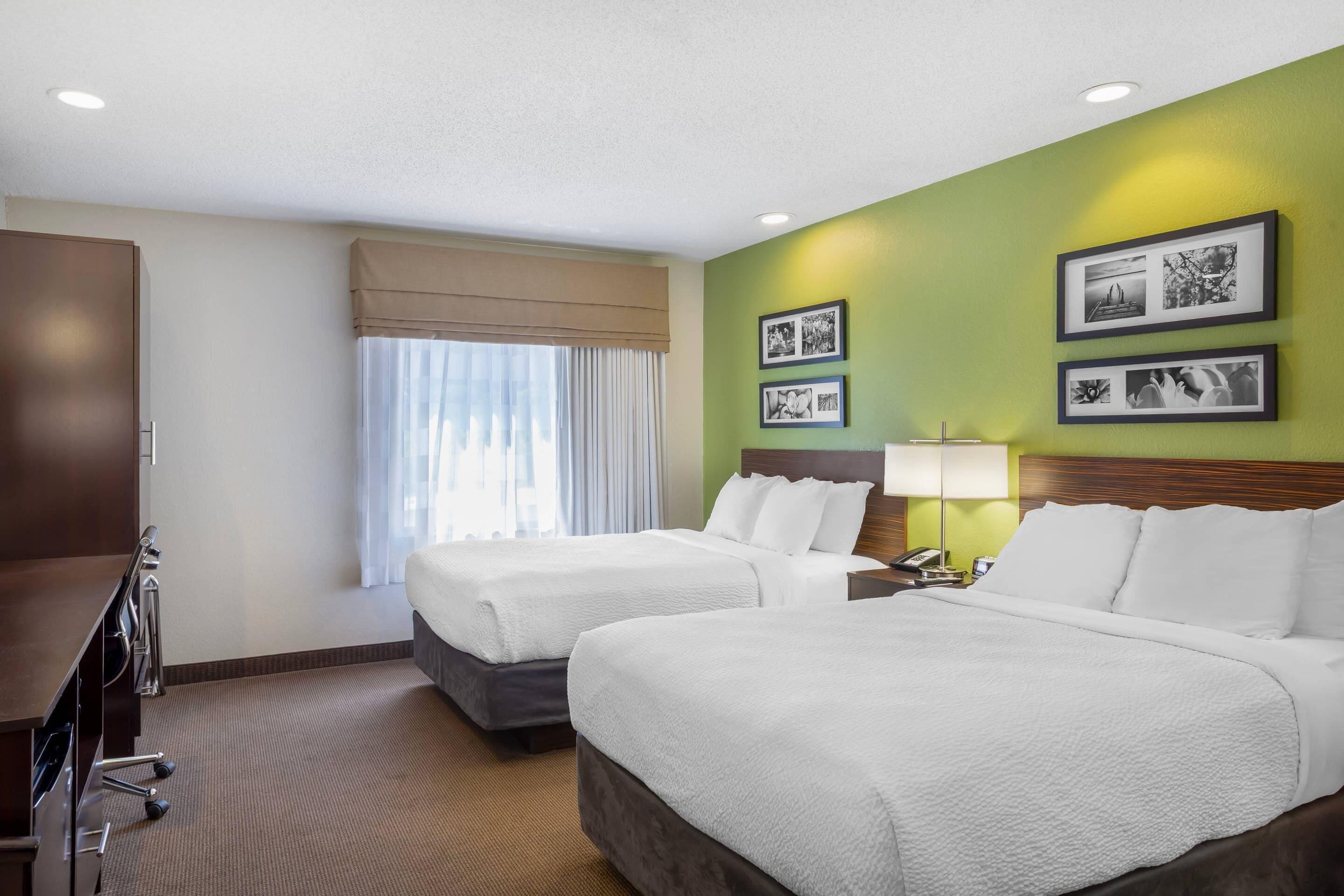 A hotel room with two beds, a desk, a lamp, and framed pictures on the wall.
