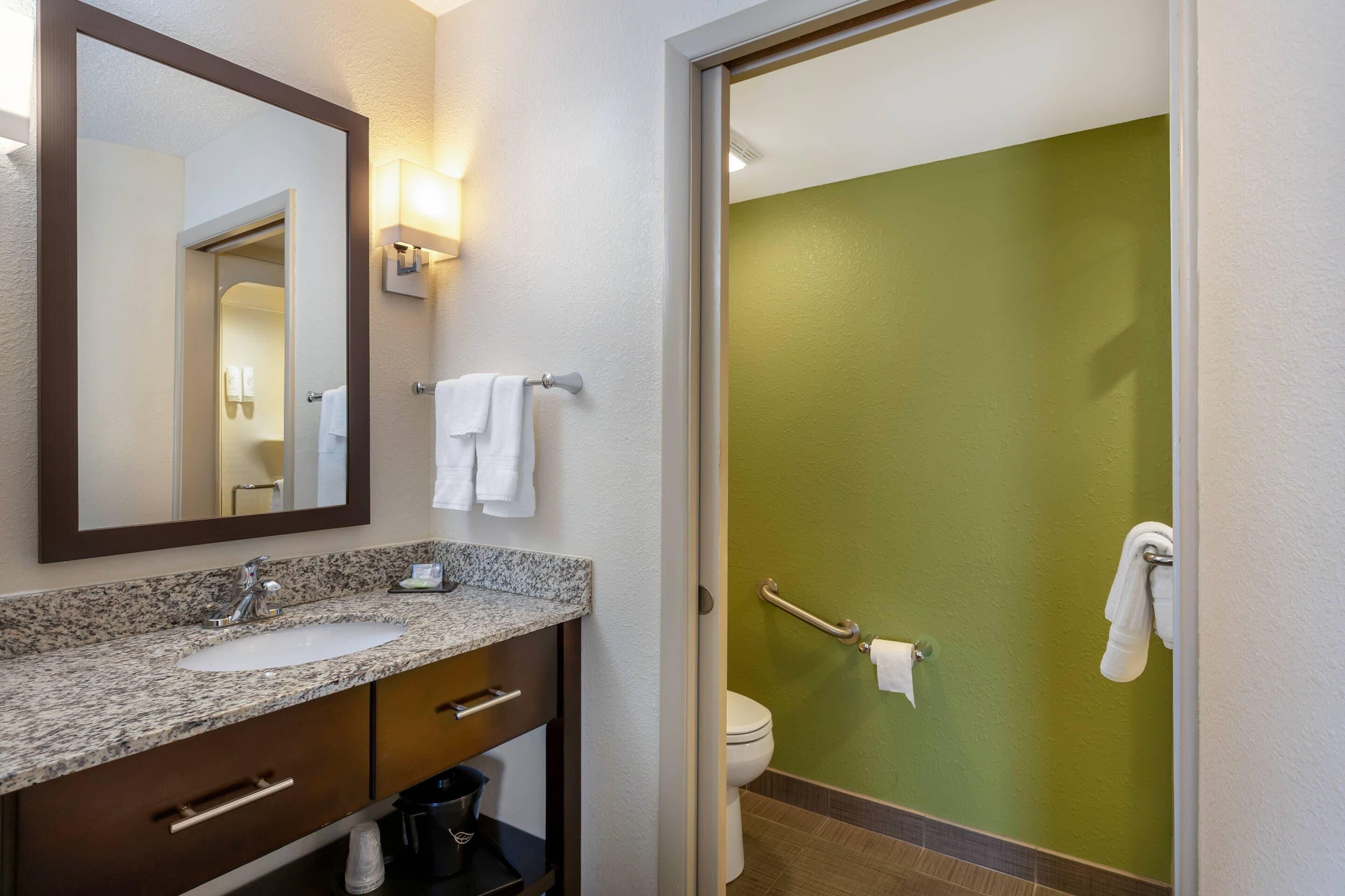A bathroom with a granite countertop sink, a mirror, a towel rack, a toilet, and a green wall.